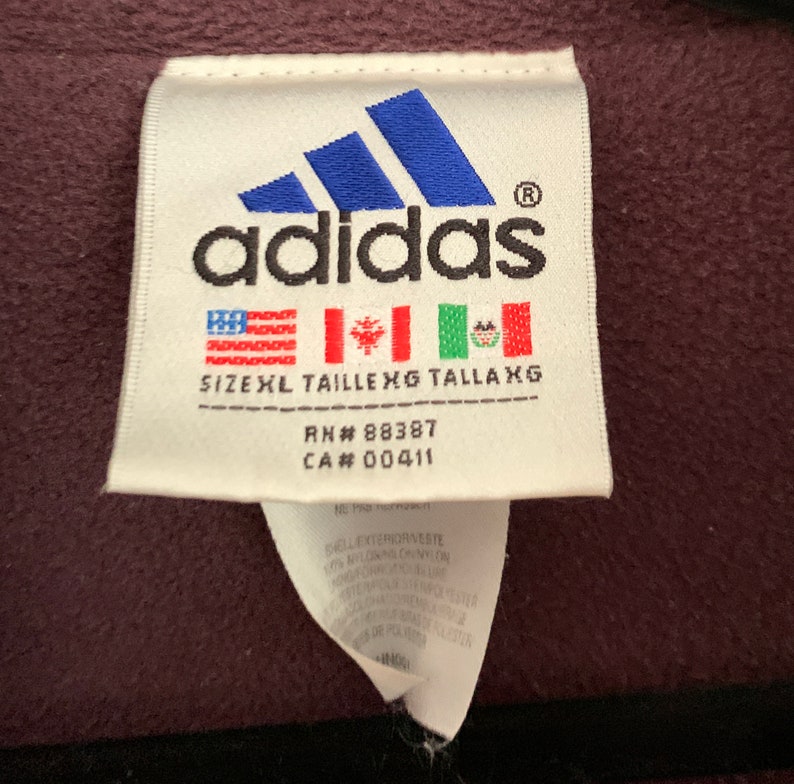 K&ouml;nnte beinhalten: Ein wei&szlig;es Etikett mit dem Adidas-Logo und dem Text "adidas" in Schwarz. Das Etikett enth&auml;lt auch den Text "SIZEXL TAILLE XG TALLAXG", "RN# 88387" und "CA# 00411". Das Etikett hat eine amerikanische Flagge in Rot, Wei&szlig; und Blau, eine kanadische Flagge und eine mexikanische Flagge.
