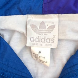 K&ouml;nnte beinhalten: Eine blau-violette Nylonjacke mit wei&szlig;em Innenfutter. Ein wei&szlig;es Etikett zeigt das Adidas-Trefoil-Logo, den Schriftzug 'adidas', 'MADE IN TAIWAN' sowie ein Etikett mit Gr&ouml;&szlig;e M, 100% Nylon-Au&szlig;enmaterial und 65% Polyester, 35% Baumwoll-Futter.