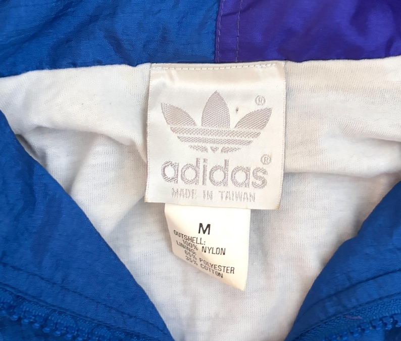 K&ouml;nnte beinhalten: Eine blau-violette Nylonjacke mit wei&szlig;em Innenfutter. Ein wei&szlig;es Etikett zeigt das Adidas-Trefoil-Logo, den Schriftzug 'adidas', 'MADE IN TAIWAN' sowie ein Etikett mit Gr&ouml;&szlig;e M, 100% Nylon-Au&szlig;enmaterial und 65% Polyester, 35% Baumwoll-Futter.