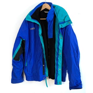 Puede incluir: Una chaqueta Columbia azul y turquesa con forro de polar negro. La chaqueta tiene cierre de cremallera y cierre de botones a presión. La chaqueta tiene capucha.