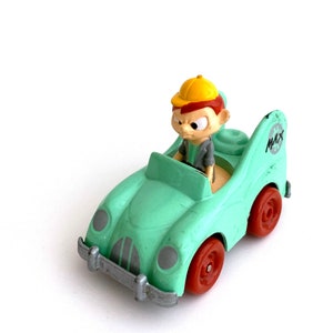 May include: A green toy car with a cartoon character driving. The car has red wheels and a silver bumper. The character is wearing a yellow hat and has a grumpy expression. The car has the word "MAX" on the side.