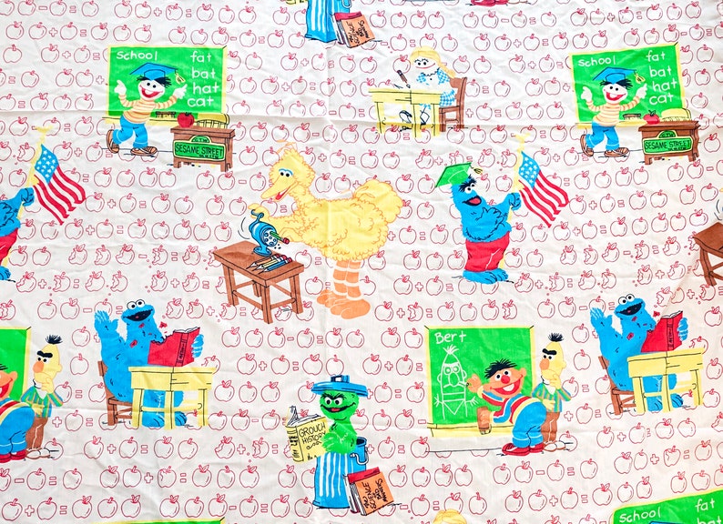 May include: A white fabric with a Sesame Street theme. The fabric features red apples, a yellow Big Bird, a blue Cookie Monster, a green Elmo, and a blue Bert. The fabric also features the text "School", "Fat", "Bat", "Hat", "Cat", "Sesame Street", and "Bert".