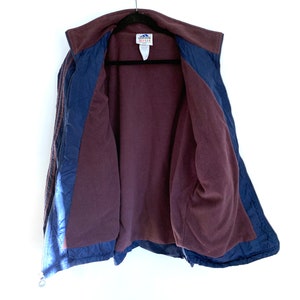 K&ouml;nnte beinhalten: Eine marineblaue und burgunderrote Fleecejacke mit Rei&szlig;verschluss und Adidas-Branding. Die Jacke hat ein gestepptes Futter und einen Stehkragen.