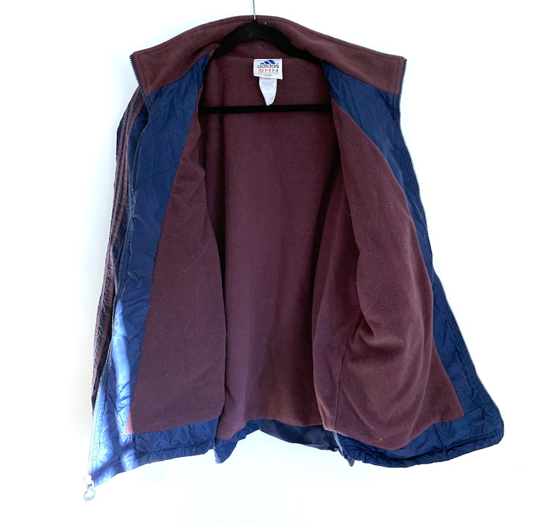 K&ouml;nnte beinhalten: Eine marineblaue und burgunderrote Fleecejacke mit Rei&szlig;verschluss und Adidas-Branding. Die Jacke hat ein gestepptes Futter und einen Stehkragen.