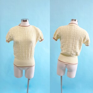 May include: Pale yellow knit top with a textured geometric pattern. The short-sleeved shirt has ribbed trim at the waist, neckline, and sleeves, accented with a thin burgundy stripe. The top is displayed on a mannequin against a light blue backdrop.