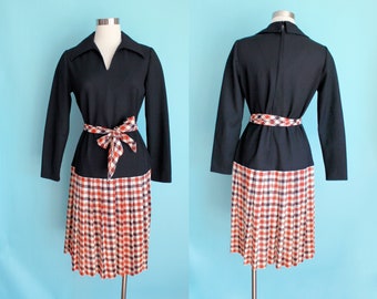 May include: A vintage black dress with long sleeves and a V-neck collar. The dress features a pleated skirt with a red, white, and black plaid pattern. A matching plaid belt cinches the waist. The dress is displayed on a mannequin.