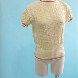 May include: A pale yellow short-sleeved sweater with a textured geometric pattern. The sweater has a ribbed hem and neckline, and the edges are trimmed with a thin burgundy stripe. The garment is displayed on a mannequin against a light blue backdrop.