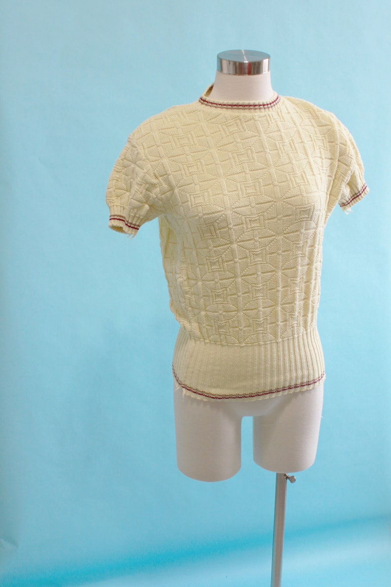 May include: A pale yellow short-sleeved sweater with a textured geometric pattern. The sweater has a ribbed hem and neckline, and the edges are trimmed with a thin burgundy stripe. The garment is displayed on a mannequin against a light blue backdrop.