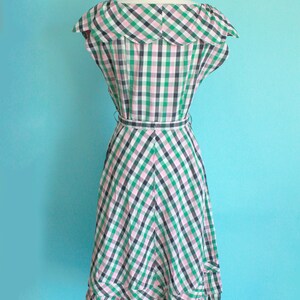 May include: A green and pink gingham dress with a ruffled collar and a fitted bodice. The dress has a full skirt with a tiered ruffle at the hem. The dress is on a mannequin against a light blue background.