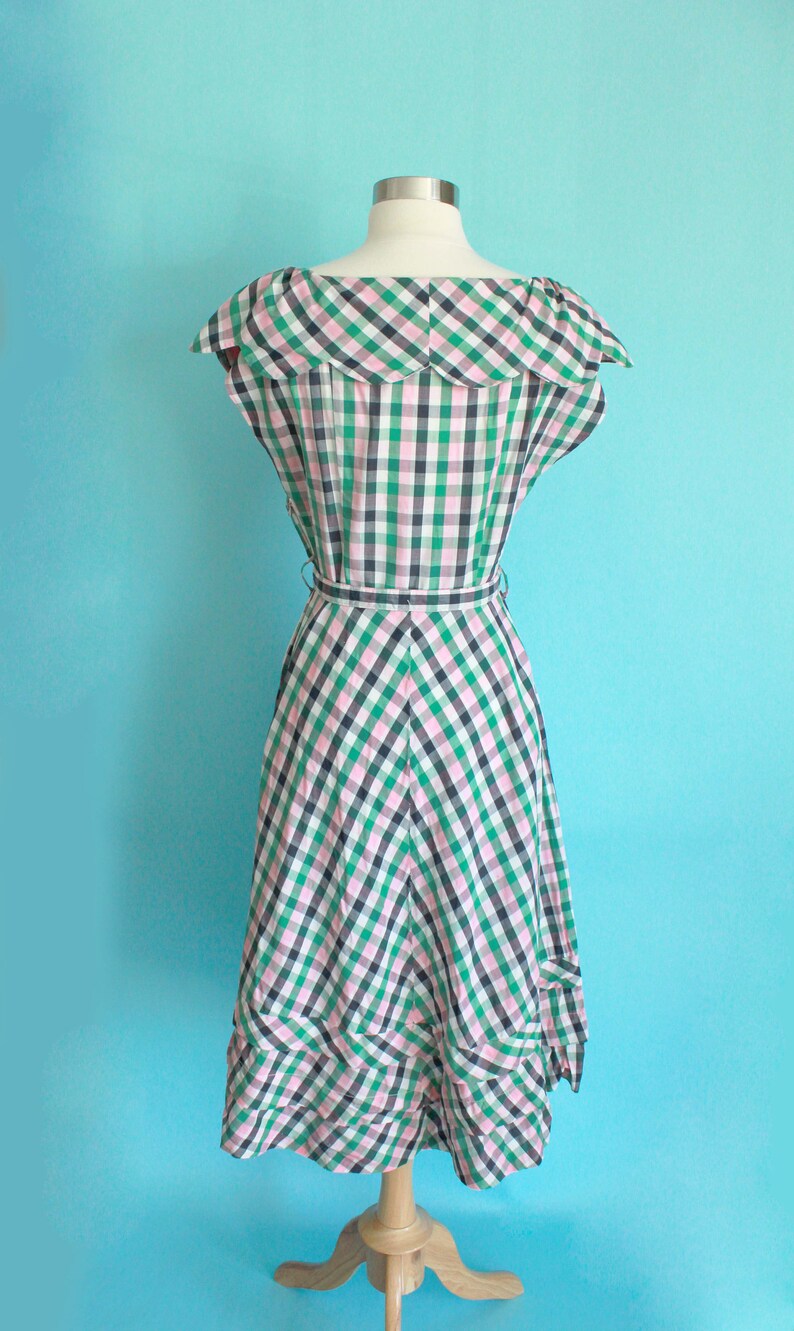 May include: A green and pink gingham dress with a ruffled collar and a fitted bodice. The dress has a full skirt with a tiered ruffle at the hem. The dress is on a mannequin against a light blue background.