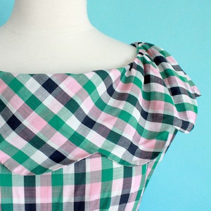 May include: A green, pink, and white plaid dress with a ruffled neckline. The dress is on a white mannequin against a light blue background.