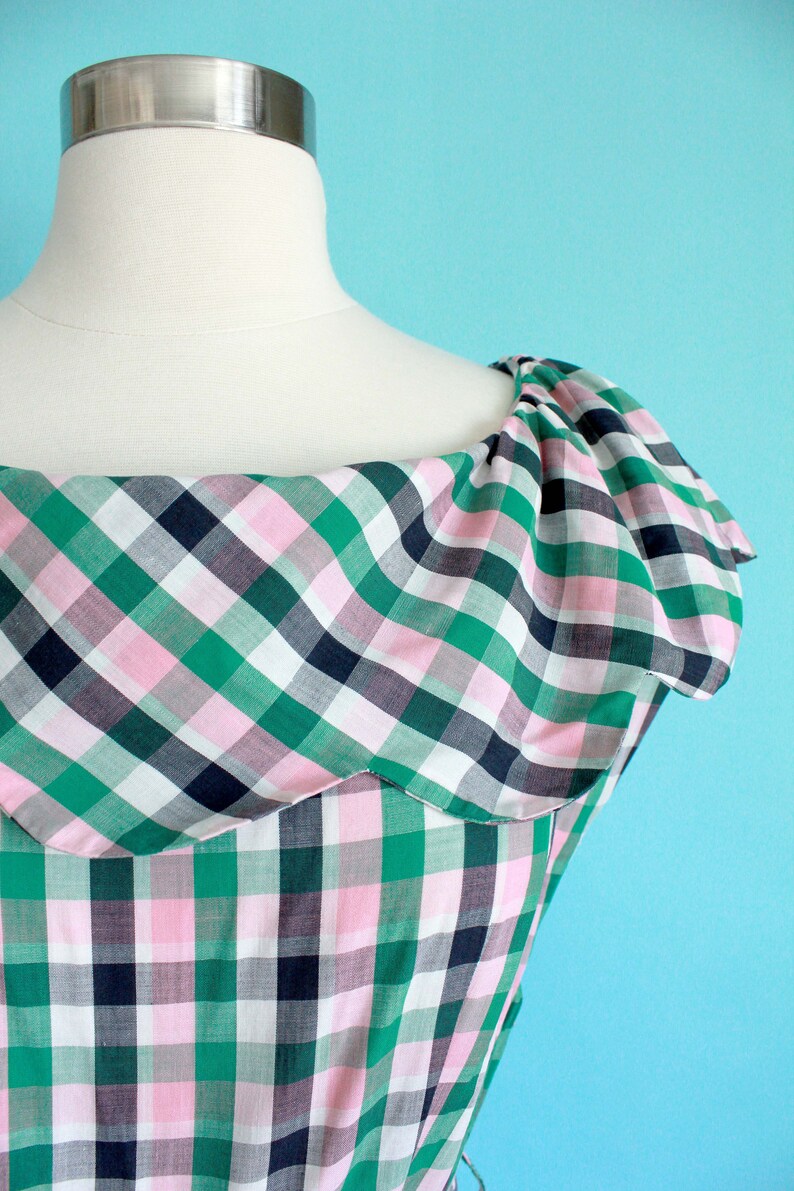 May include: A green, pink, and white plaid dress with a ruffled neckline. The dress is on a white mannequin against a light blue background.