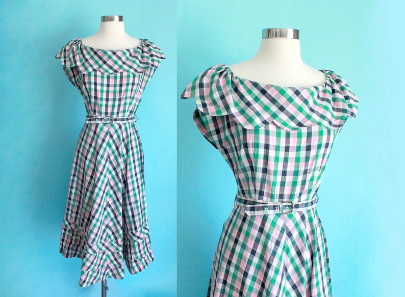 May include: A green, pink, and black gingham dress with a ruffled neckline and a white belt. The dress has a fitted bodice and a full skirt.
