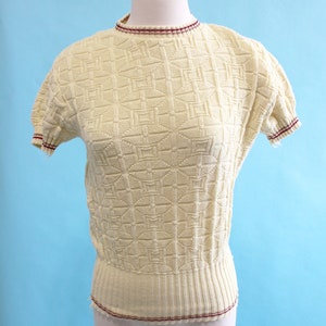 May include: Cream-colored vintage knit top with a geometric pattern. The short-sleeved top features a ribbed waist and neckline and sleeve trim with a burgundy stripe. The top is displayed on a mannequin against a light blue background.
