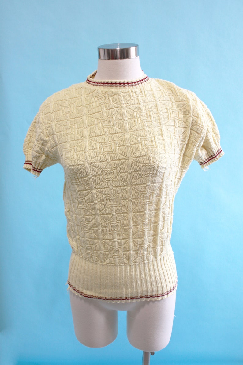 May include: Cream-colored vintage knit top with a geometric pattern. The short-sleeved top features a ribbed waist and neckline and sleeve trim with a burgundy stripe. The top is displayed on a mannequin against a light blue background.