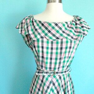 May include: A green, pink, and black gingham dress with a ruffled neckline and a belt. The dress is on a white mannequin against a light blue background.