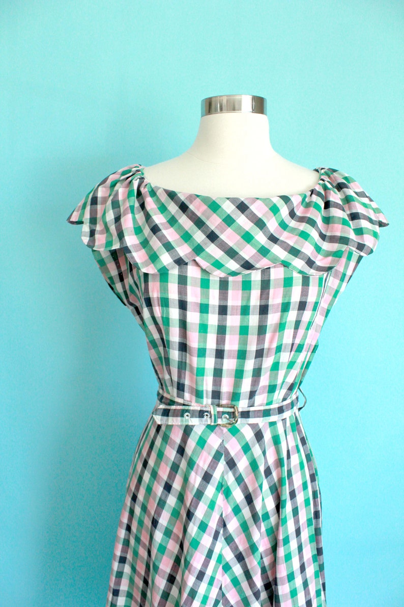 May include: A green, pink, and black gingham dress with a ruffled neckline and a belt. The dress is on a white mannequin against a light blue background.