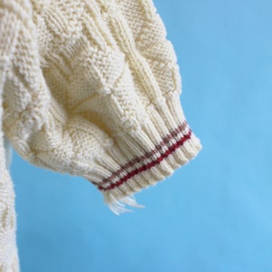 May include: Close-up of a cream-colored knitted sweater sleeve. The sleeve features a cable knit pattern and a ribbed cuff with burgundy and brown stripes. Frayed threads hang from the cuff against a blue background.