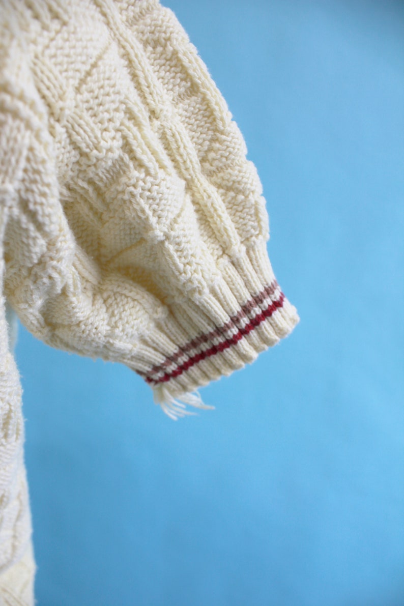 May include: Close-up of a cream-colored knitted sweater sleeve. The sleeve features a cable knit pattern and a ribbed cuff with burgundy and brown stripes. Frayed threads hang from the cuff against a blue background.