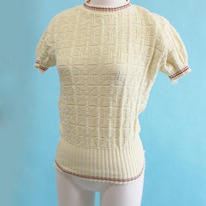 May include: Cream-colored knit top with a geometric pattern. The short-sleeved sweater has a ribbed hem and neckline, both accented with a thin burgundy stripe. The top is displayed on a white mannequin against a light blue backdrop.