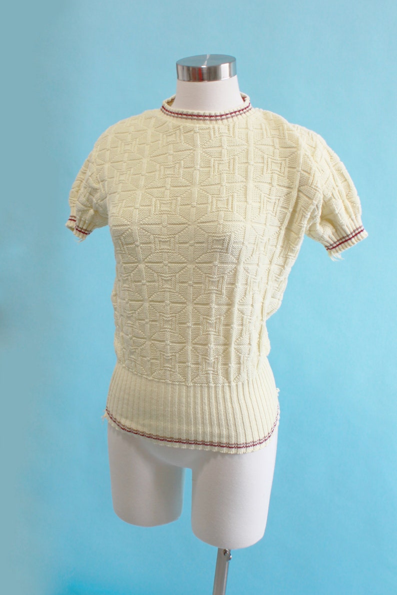 May include: Cream-colored knit top with a geometric pattern. The short-sleeved sweater has a ribbed hem and neckline, both accented with a thin burgundy stripe. The top is displayed on a white mannequin against a light blue backdrop.