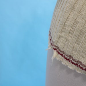 May include: Close-up of a cream-colored, ribbed knit fabric with a burgundy and brown stitched edge. The fabric appears to be part of a garment, possibly a sweater or top, against a light blue background.