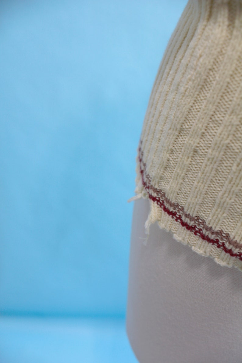 May include: Close-up of a cream-colored, ribbed knit fabric with a burgundy and brown stitched edge. The fabric appears to be part of a garment, possibly a sweater or top, against a light blue background.