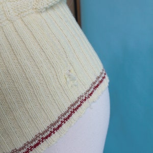 May include: Close-up of a cream-colored knitted sweater. The sweater has a ribbed design with a decorative pattern on the upper part. The bottom edge features a red and brown stripe detail. There is a small hole in the fabric.