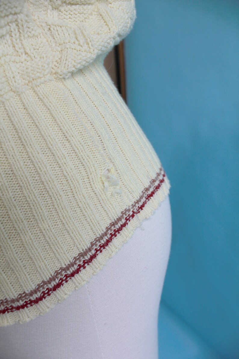 May include: Close-up of a cream-colored knitted sweater. The sweater has a ribbed design with a decorative pattern on the upper part. The bottom edge features a red and brown stripe detail. There is a small hole in the fabric.