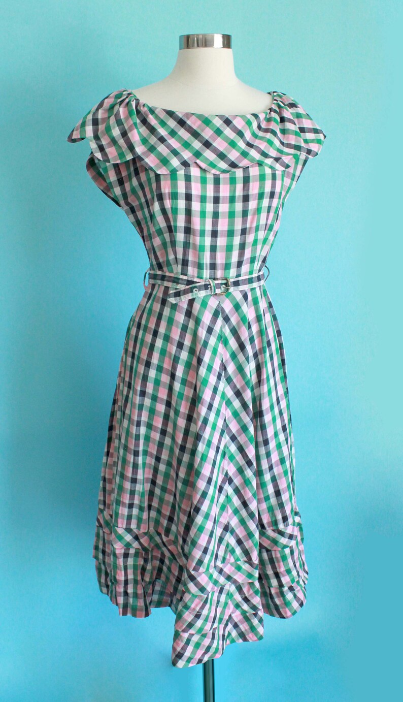 May include: A green, pink, and black gingham dress with a ruffled neckline and a matching belt. The dress has a full skirt and a scalloped hem.