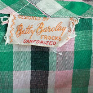 May include: A white tag with orange text that reads "Designed by Betty Barclay Frocks Sanforized". The tag is attached to a green and pink gingham fabric.