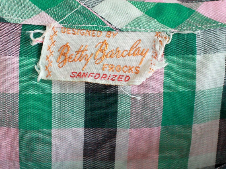 May include: A white tag with orange text that reads "Designed by Betty Barclay Frocks Sanforized". The tag is attached to a green and pink gingham fabric.
