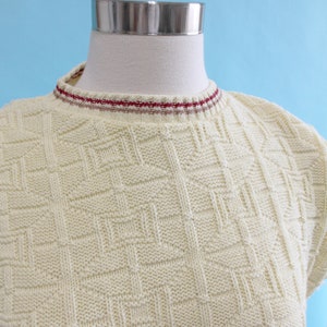 May include: Cream-colored knit top with a geometric pattern. The top has a crew neck with a red and white striped trim. The short-sleeved top is displayed on a mannequin against a light blue background.