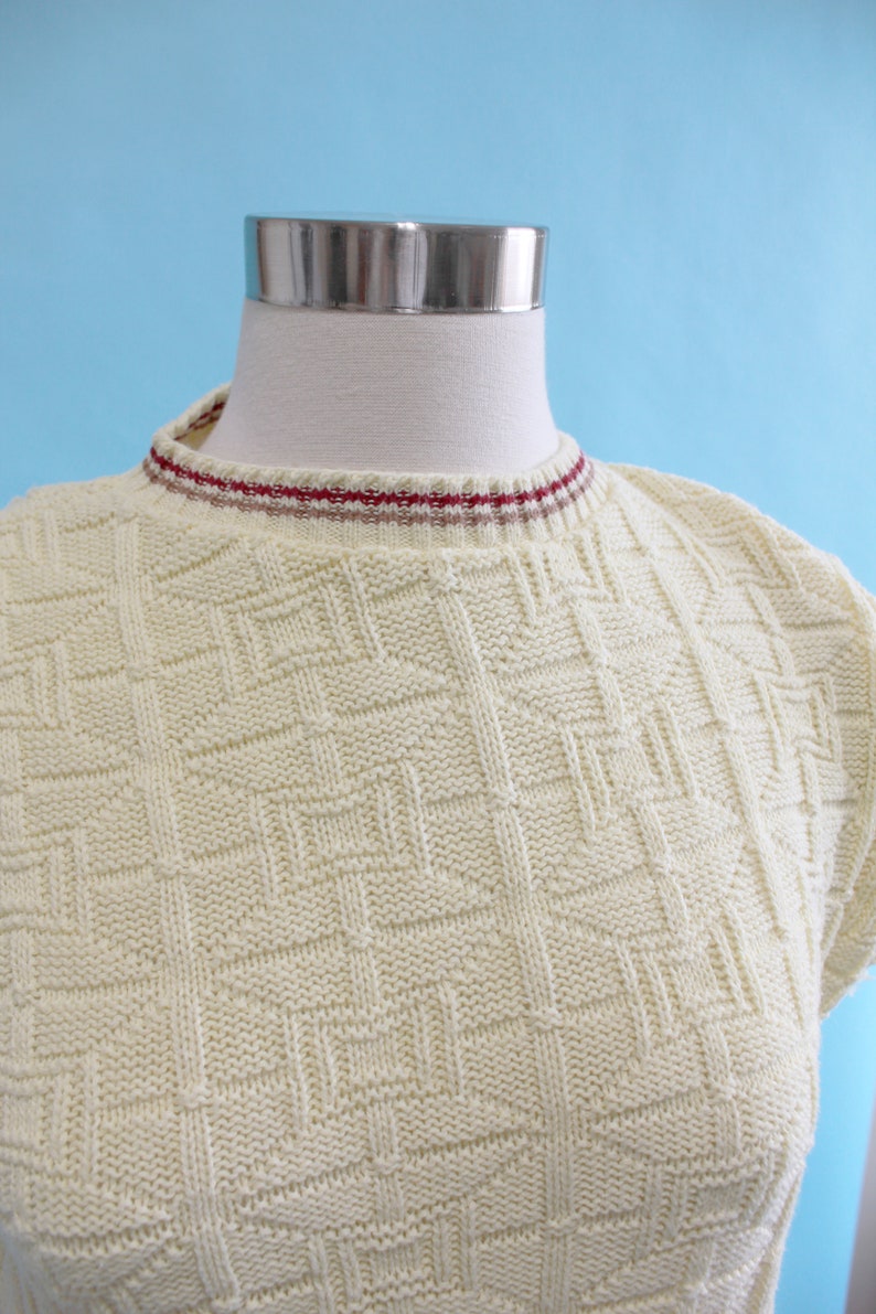 May include: Cream-colored knit top with a geometric pattern. The top has a crew neck with a red and white striped trim. The short-sleeved top is displayed on a mannequin against a light blue background.