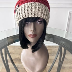 May include: A handmade crochet beanie in beige, red, and yellow. The hat features a wide red stripe and a small metal tag. The beanie is displayed on a mannequin head with black hair.