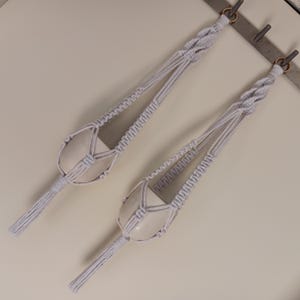 May include: Two white macrame plant hangers with tassels hanging from a wall.