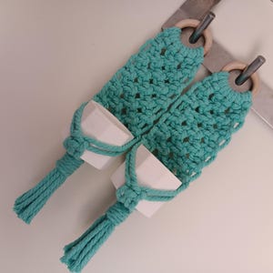 May include: Two turquoise macrame plant hangers with white ceramic pots. Each hanger has a wooden ring at the top and a knotted design. These hangers are designed to hold small plants or flowers, adding a touch of bohemian style to any space.
