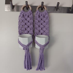 May include: Two purple macrame plant hangers with white ceramic pots. The hangers are attached to a metal bar with wooden rings.