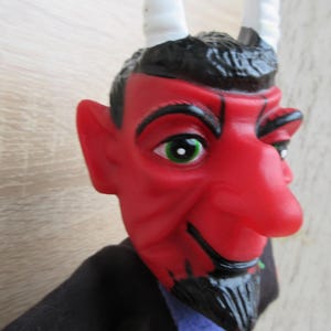 Könnte beinhalten: Eine rote Teufelspuppe mit weißen Hörnern, schwarzen Haaren und einem schwarzen Ziegenbart. Die Puppe hat eine große, rote Nase und grüne Augen.