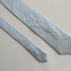 May include: A light blue necktie with a repeating diamond pattern in white and light blue. The tie is made of a smooth fabric and is displayed on a white surface. The tie is a classic accessory for formal or business attire.