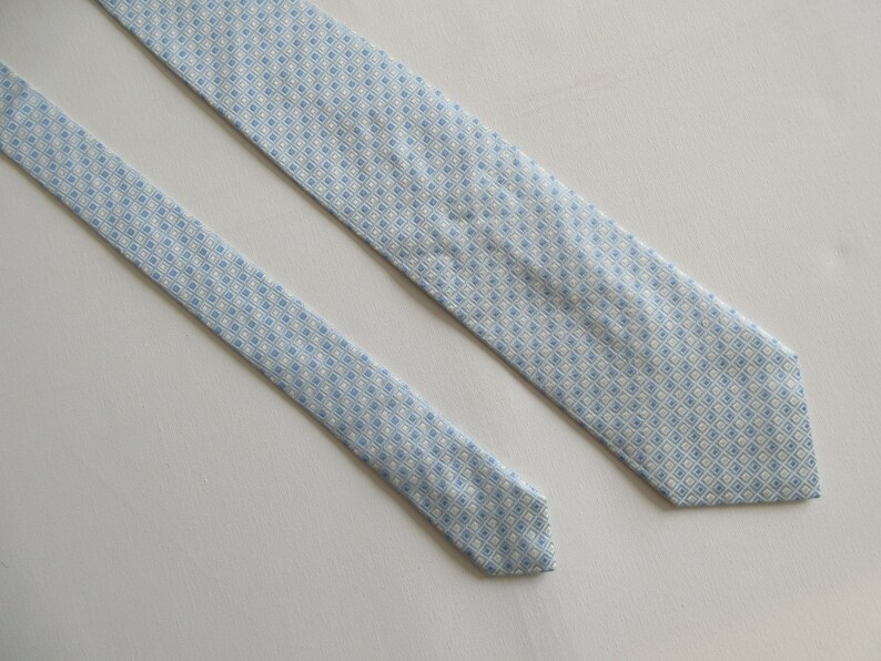 May include: A light blue necktie with a repeating diamond pattern in white and light blue. The tie is made of a smooth fabric and is displayed on a white surface. The tie is a classic accessory for formal or business attire.