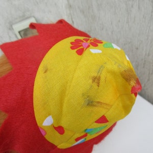 May include: A bright red felt hat featuring a yellow fabric crown with a floral pattern. The yellow section displays small red, green, and white petal shapes, with visible brown stains on the fabric surface near the top point.