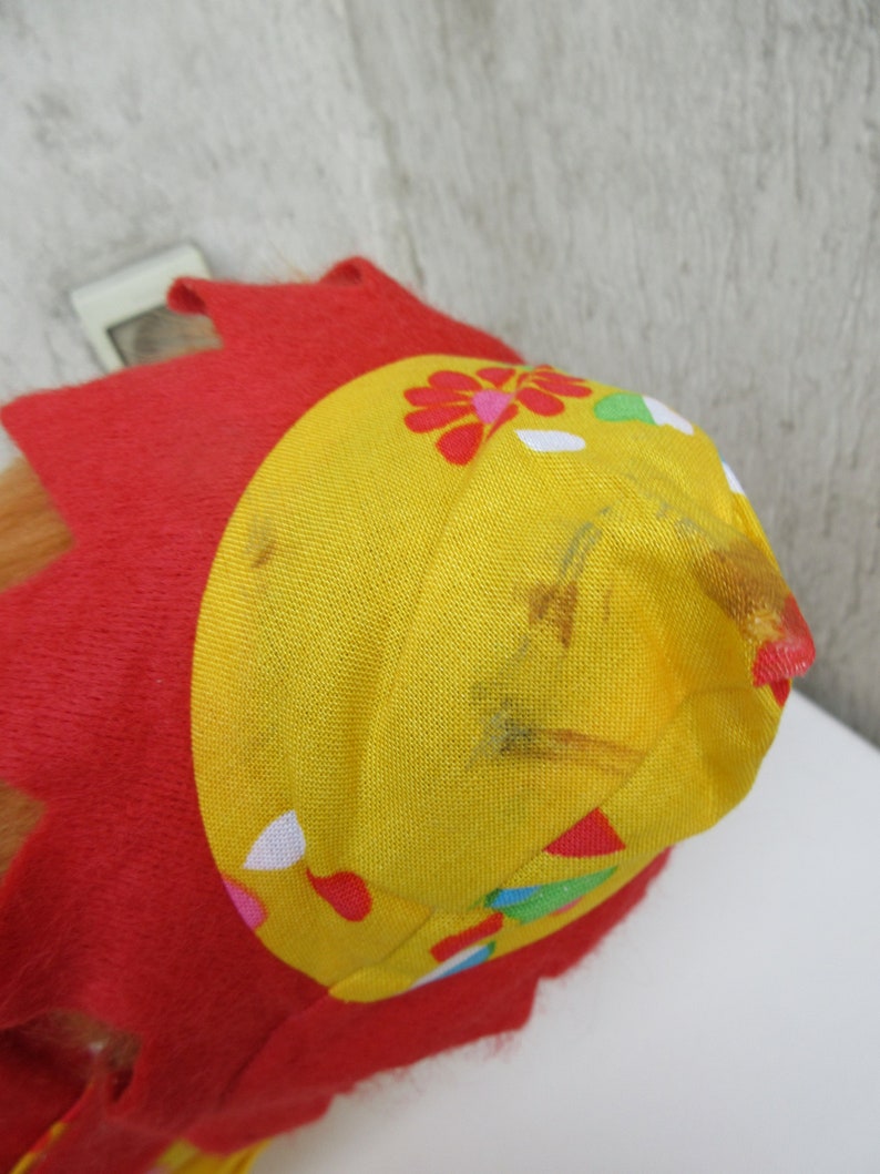 May include: A bright red felt hat featuring a yellow fabric crown with a floral pattern. The yellow section displays small red, green, and white petal shapes, with visible brown stains on the fabric surface near the top point.