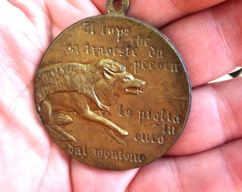Puede incluir: Un medallón circular de metal en tono bronce con un relieve de un lobo vestido con piel de oveja. El texto italiano "Il lupo che si traveste da pecora lo piglia in culo dal montone" está grabado alrededor de la figura central.