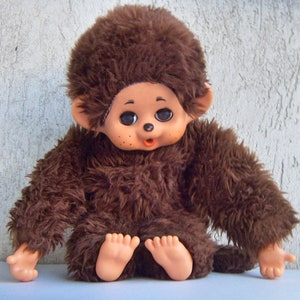 May include: A brown plush monkey toy with large black eyes and a pink nose. The monkey is sitting with its legs crossed and its arms outstretched.