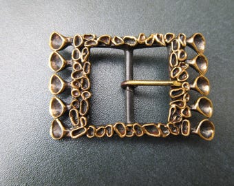 May include: A rectangular belt buckle with a dark bronze finish, featuring a textured, organic, cellular pattern along the frame and five protruding, cup-shaped elements on each side, complete with a central metal prong for fastening.