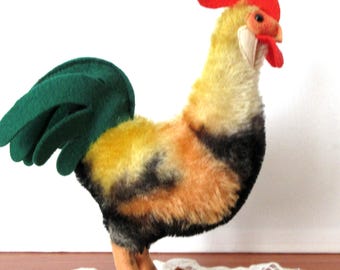 May include: A plush rooster figurine featuring a bright red felt comb and wattle, a yellow and orange fuzzy body with black accents, and a dark green felt tail. The bird stands on two light brown felt legs with defined feet.