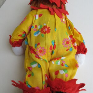 May include: A clown doll wearing a yellow jumpsuit with a colourful floral pattern, red ruffled trim at the collar, wrists, and ankles, a matching yellow pointed hat with red trim, white fabric hands, and black fabric shoes.