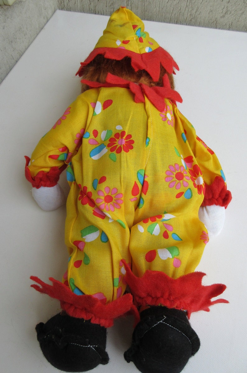 May include: A clown doll wearing a yellow jumpsuit with a colourful floral pattern, red ruffled trim at the collar, wrists, and ankles, a matching yellow pointed hat with red trim, white fabric hands, and black fabric shoes.