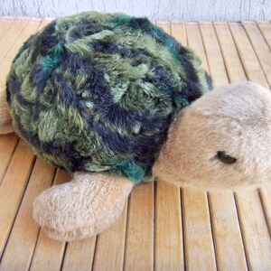 May include: A soft toy turtle featuring a shell with a camouflage pattern in green and navy. The turtle has tan flippers, head, and tail. The toy is designed to look like a turtle and is placed on a wooden surface.